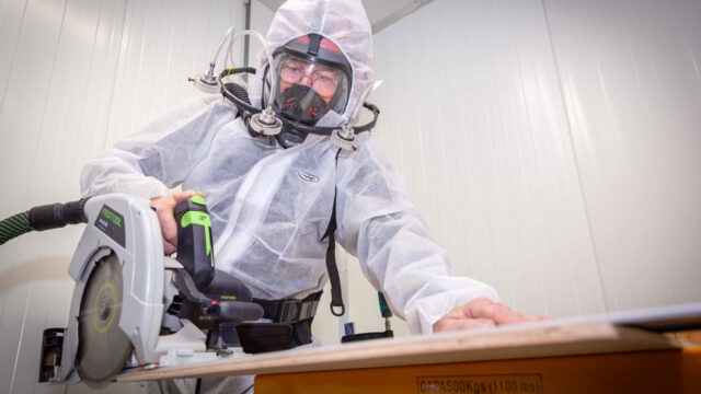 Medewerker in beschermpak en masker zaagt hout, illustratie van stofvrij werken en blootstellingsbeperking op de werkvloer