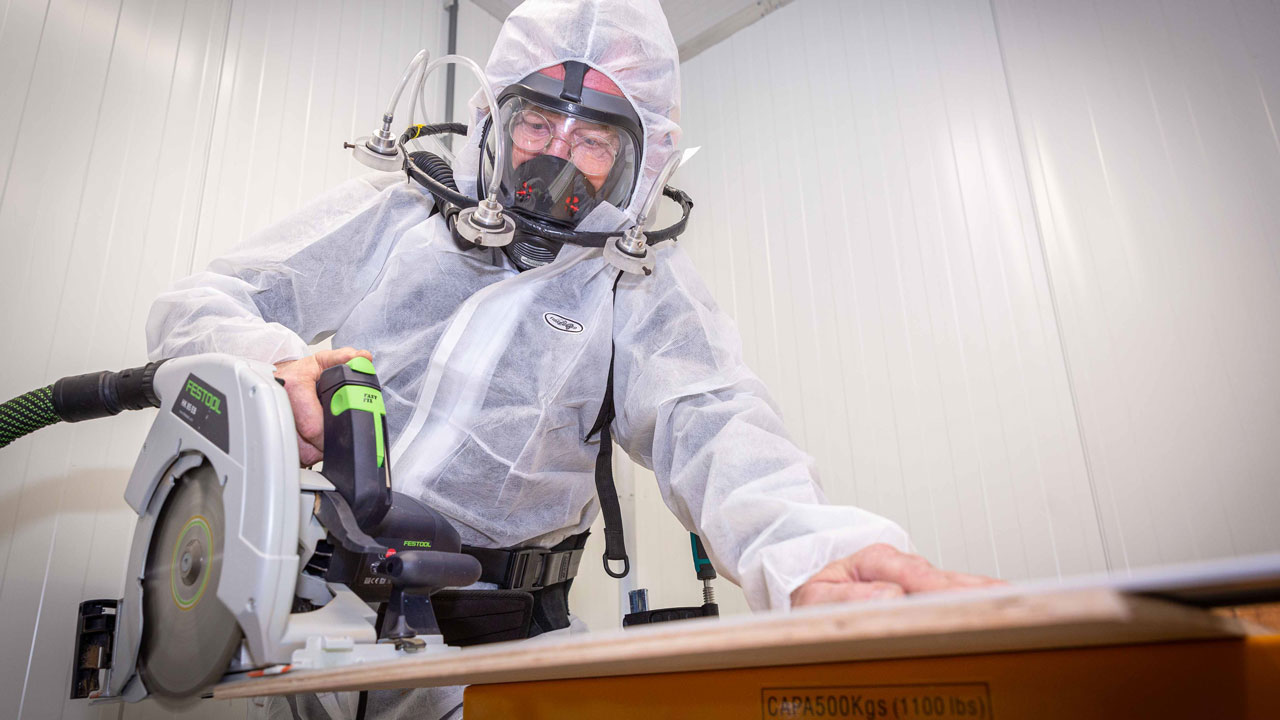 Medewerker in beschermpak en masker zaagt hout, illustratie van stofvrij werken en blootstellingsbeperking op de werkvloer