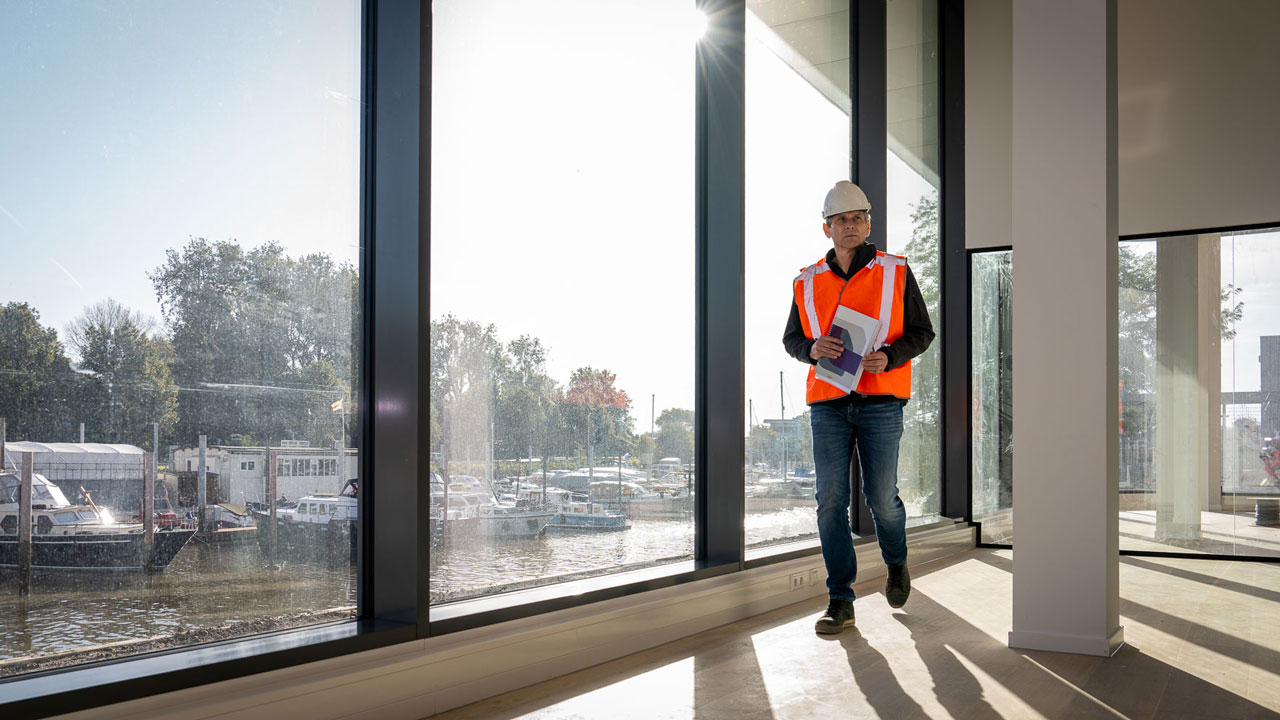 Medewerker met bouwtekeningen in duurzaam gebouw met uitzicht op jachthaven, illustratie van duurzaam bouwen in Schiedam