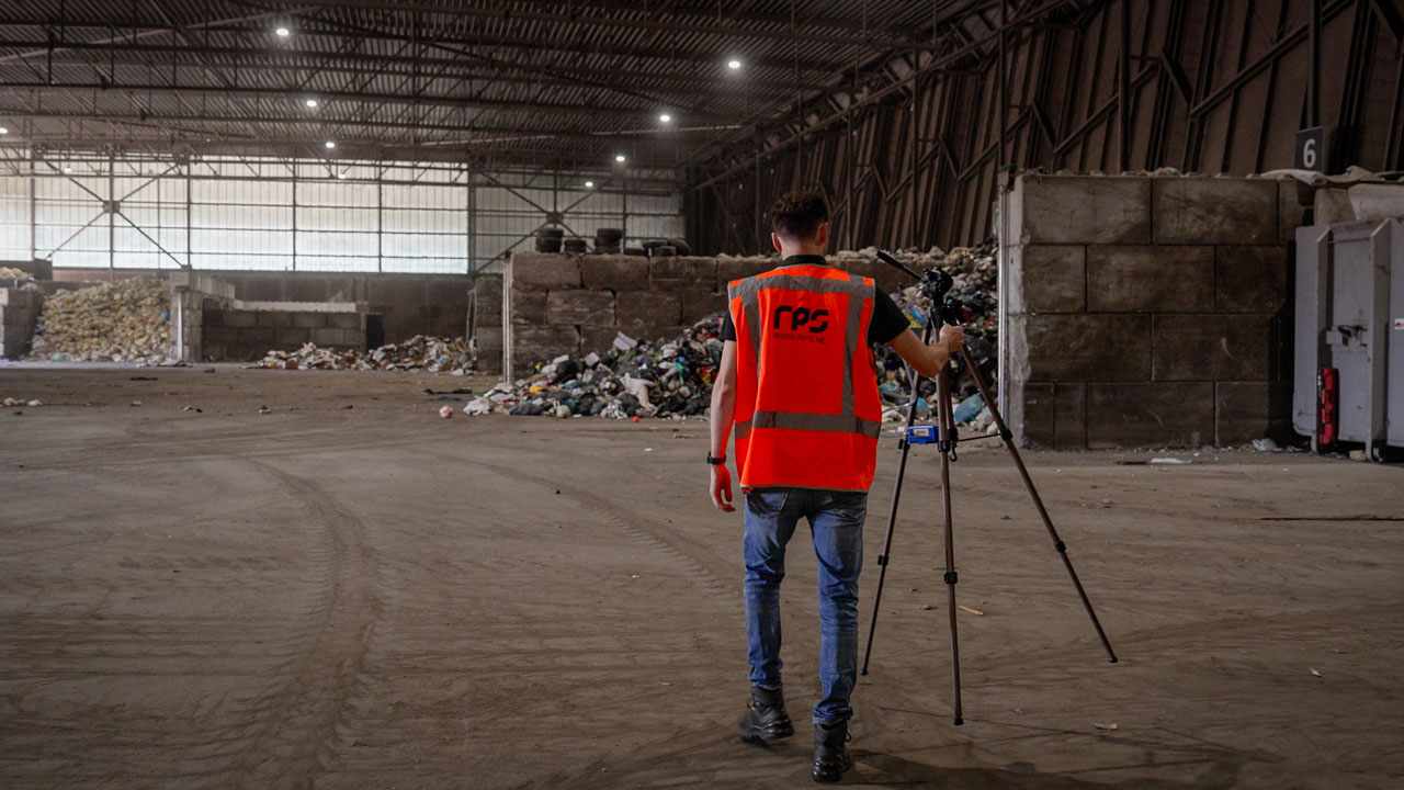 Arbeidshygiënist op vuilstort die loopt met een apparaat voor luchtmetingen