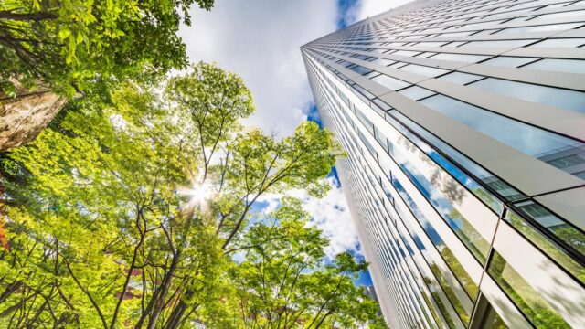 Modern hoog gebouw met glazen gevel, omringd door groene bomen en zonlicht; focus op energie-efficiëntie en duurzaam vastgoedbeheer