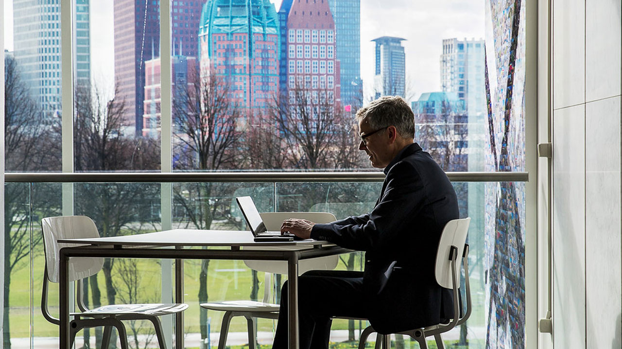 Persoon met laptop aan tafel in lichte ruimte met uitzicht op stedelijke skyline; focus op digitale verwerking van energielabeldata