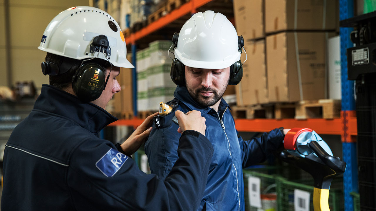 Arbeidshygiënist plaatst geluidsmeetapparatuur op schouder van medewerker voor meting blootstelling geluid