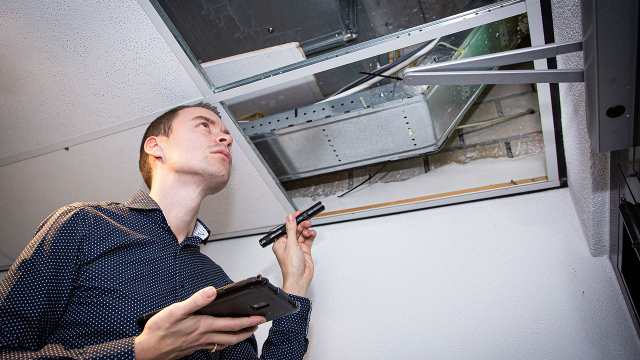 Brandveiligheidsadviseur inspecteert plafond met zaklamp en tablet bij open paneel met leidingen en bekabeling