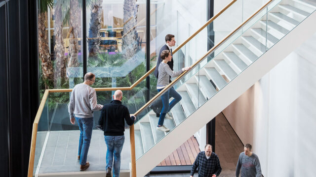 Moderne kantoorruimte met glazen gevel en grote trap, verlicht door natuurlijk licht. Mensen bewegen zich door de ruimte, met zicht op palmbomen buiten, wat een open en professionele sfeer uitstraalt