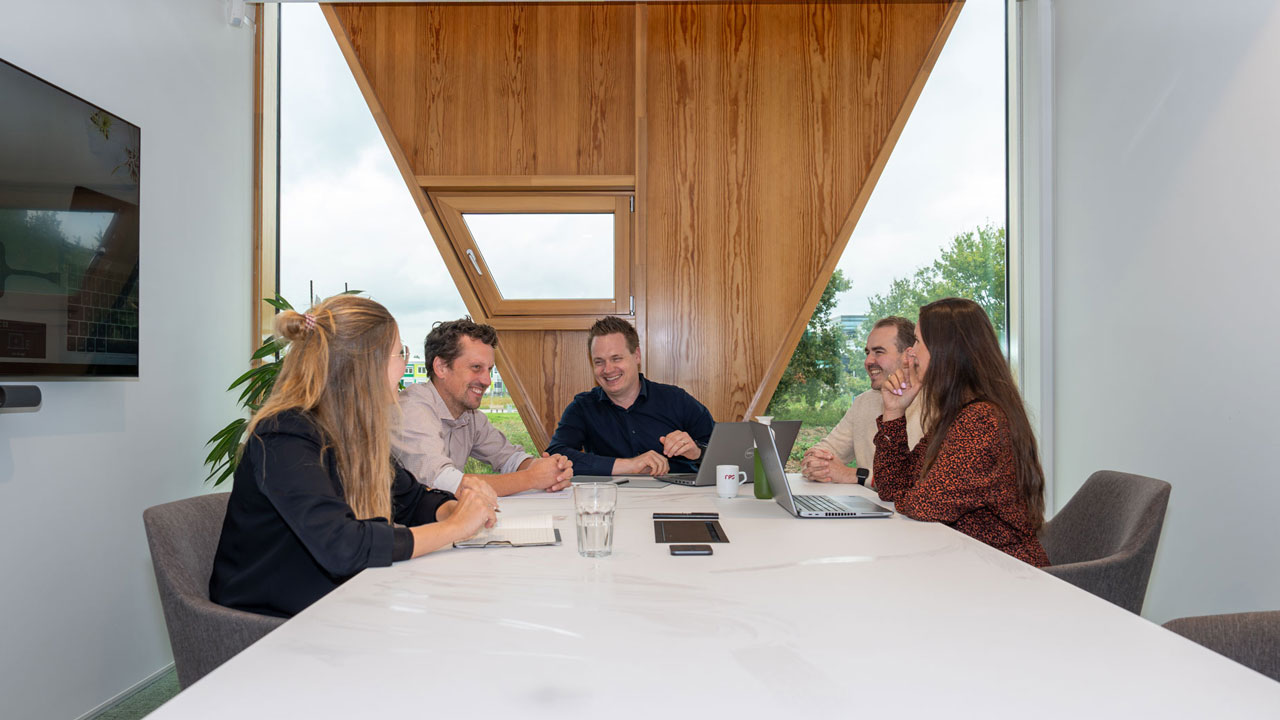 Vastgoedconsultants in overleg aan een vergadertafel in een moderne ruimte met grote ramen, laptops en notitieblokken op tafel