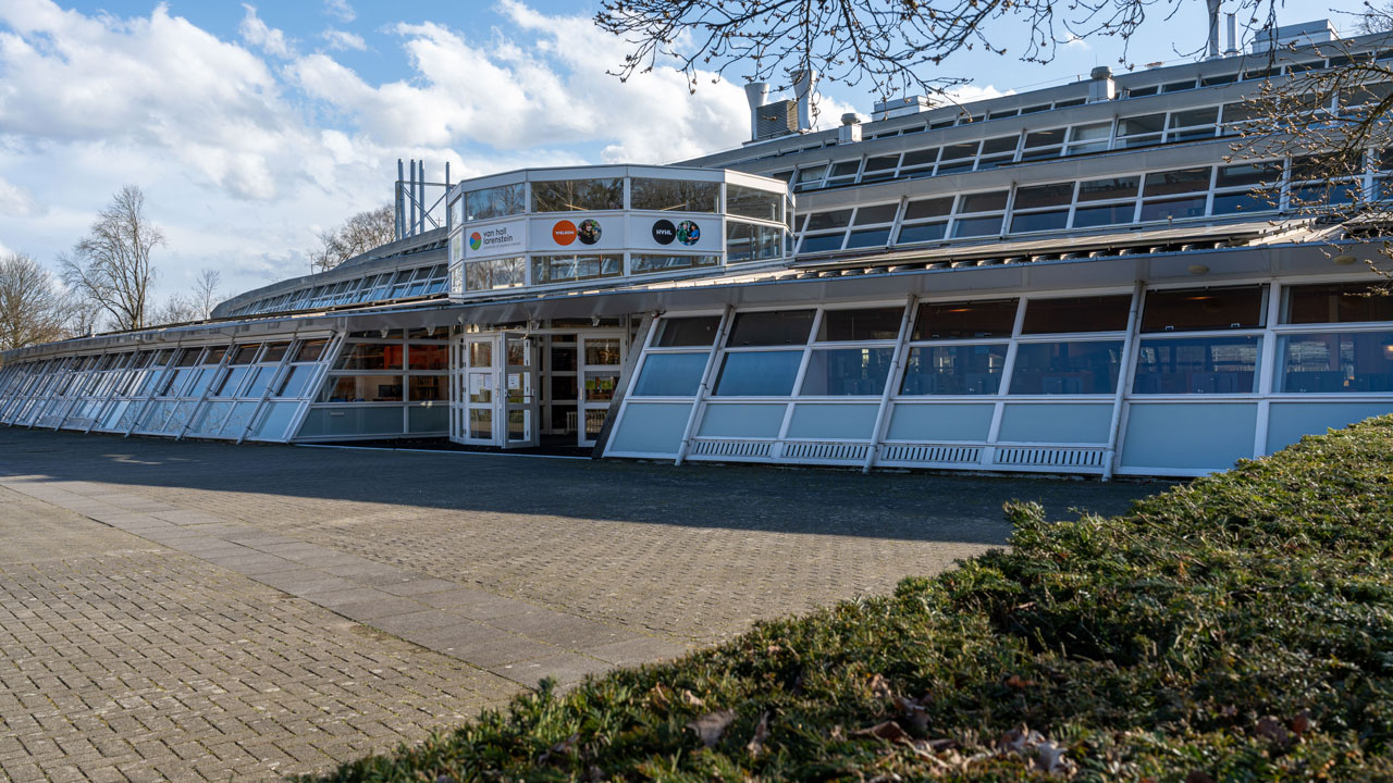 Modern schoolgebouw met gebogen gevel en grote ramen, centrale entree met glasdeuren en logo’s, omgeven door groen en bestrating