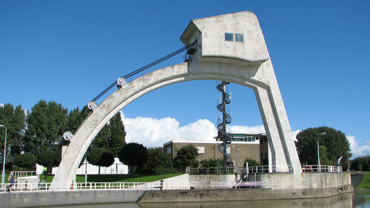 Grote betonnen boogconstructie met spiraaltrap, onderdeel van een waterbouwkundig systeem zoals een sluis of stuw, met kabels en een bedieningsgebouw bovenop