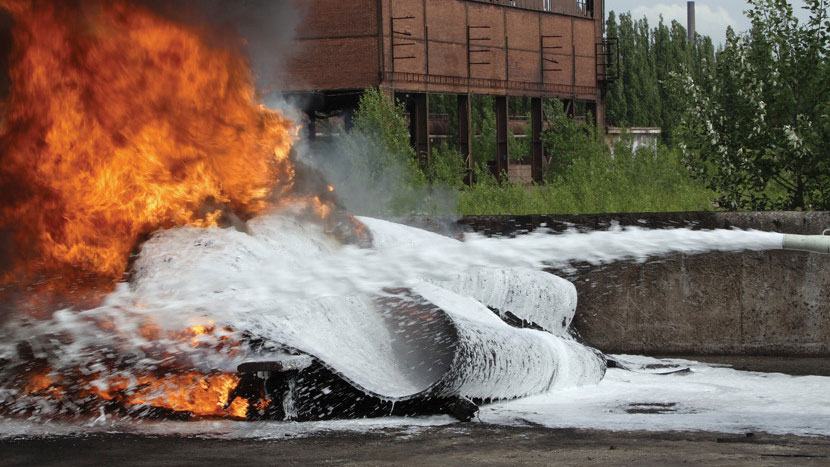 Een industriële brand wordt geblust met schuim, waarbij een gebogen metalen oppervlak en een bakstenen gebouw op de achtergrond zichtbaar zijn — verwijzend naar het gebruik van PFAS-houdend blusschuim