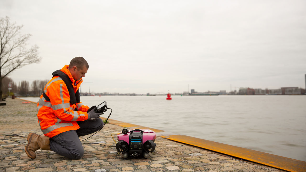 Operator in veiligheidsjas bedient roze ROV-onderwaterdrone aan waterkant – inzet van drones voor inspectie van onderwaterconstructies