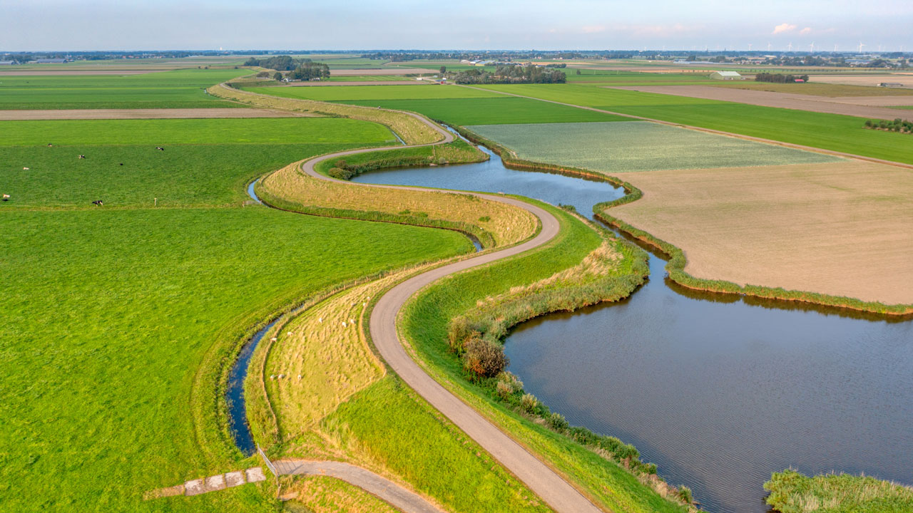 Luchtfoto van een kronkelend kanaal tussen groene en bruine landbouwvelden, met een smalle weg langs de oever en een heldere blauwe lucht erboven