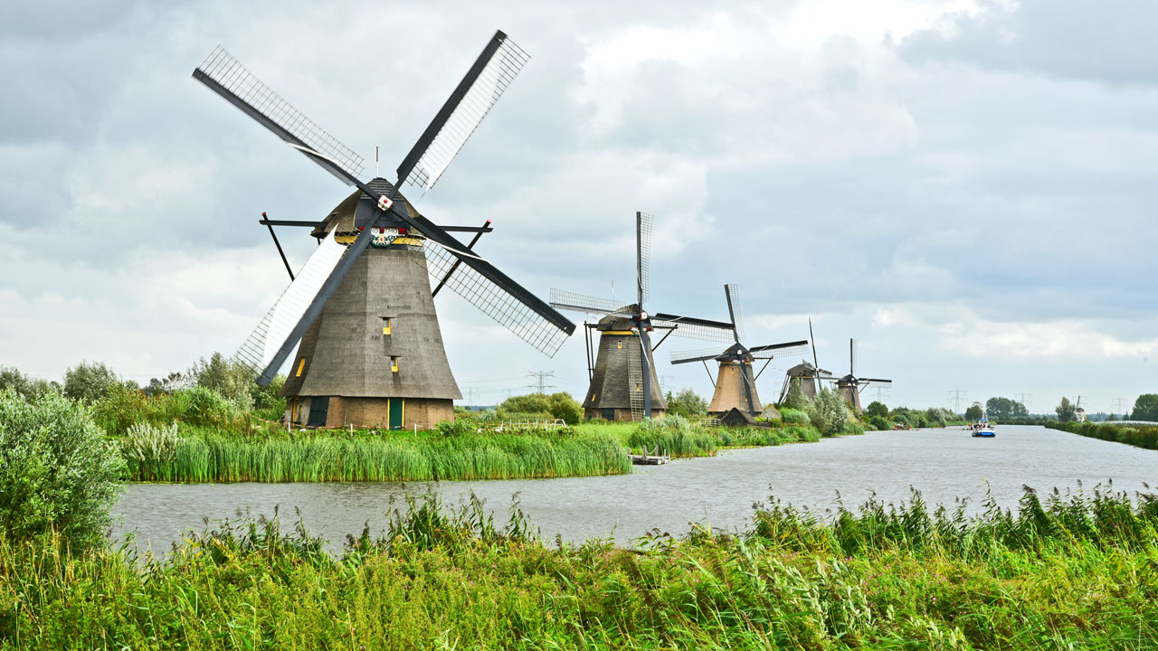 Traditionele Nederlandse windmolens langs een watergang, met groene oevers op de voorgrond en een bewolkte lucht erboven — symbool voor het samenspel tussen waterbeheer en bodemkwaliteit