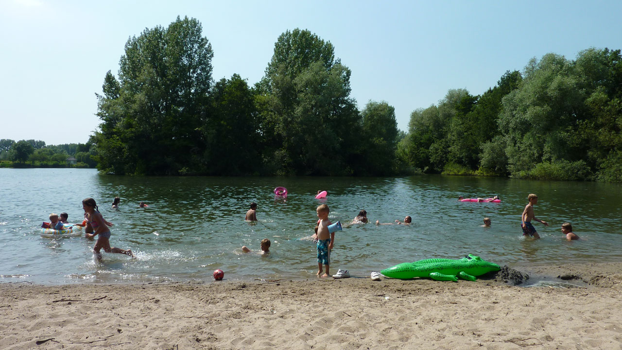 Kinderen en volwassenen zwemmen en spelen met opblaasspeelgoed in een meer naast een zandstrand, omringd door groene bomen en een blauwe lucht