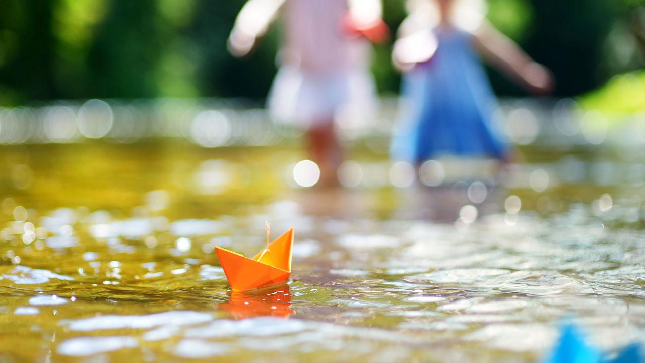 Een oranje papieren bootje drijft op helder, ondiep water met twee onscherpe kinderen en groene natuur op de achtergrond