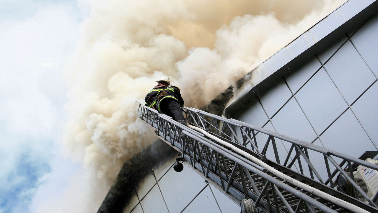 Brandweerman beklimt een uitschuifbare ladder richting het dak van een modern gebouw met glasgevel, waar rook uit opstijgt tijdens een brandincident