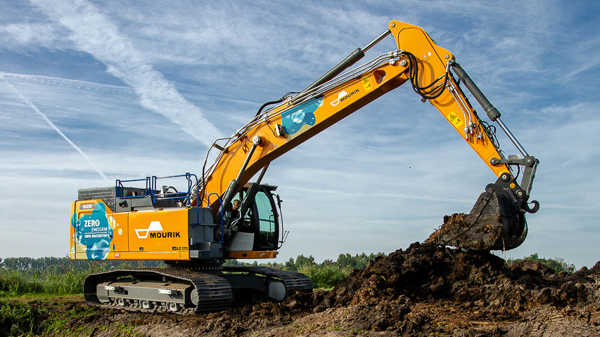 Graafmachine voert grondwerk uit langs waterkant, onderdeel van dijkversterking en waterveiligheidsproject