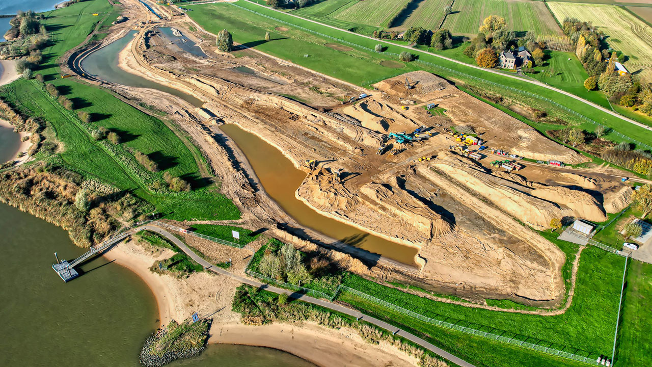 Luchtfoto van dijkverbeteringsproject met grondverzet en machines langs water, gericht op versterking en waterveiligheid