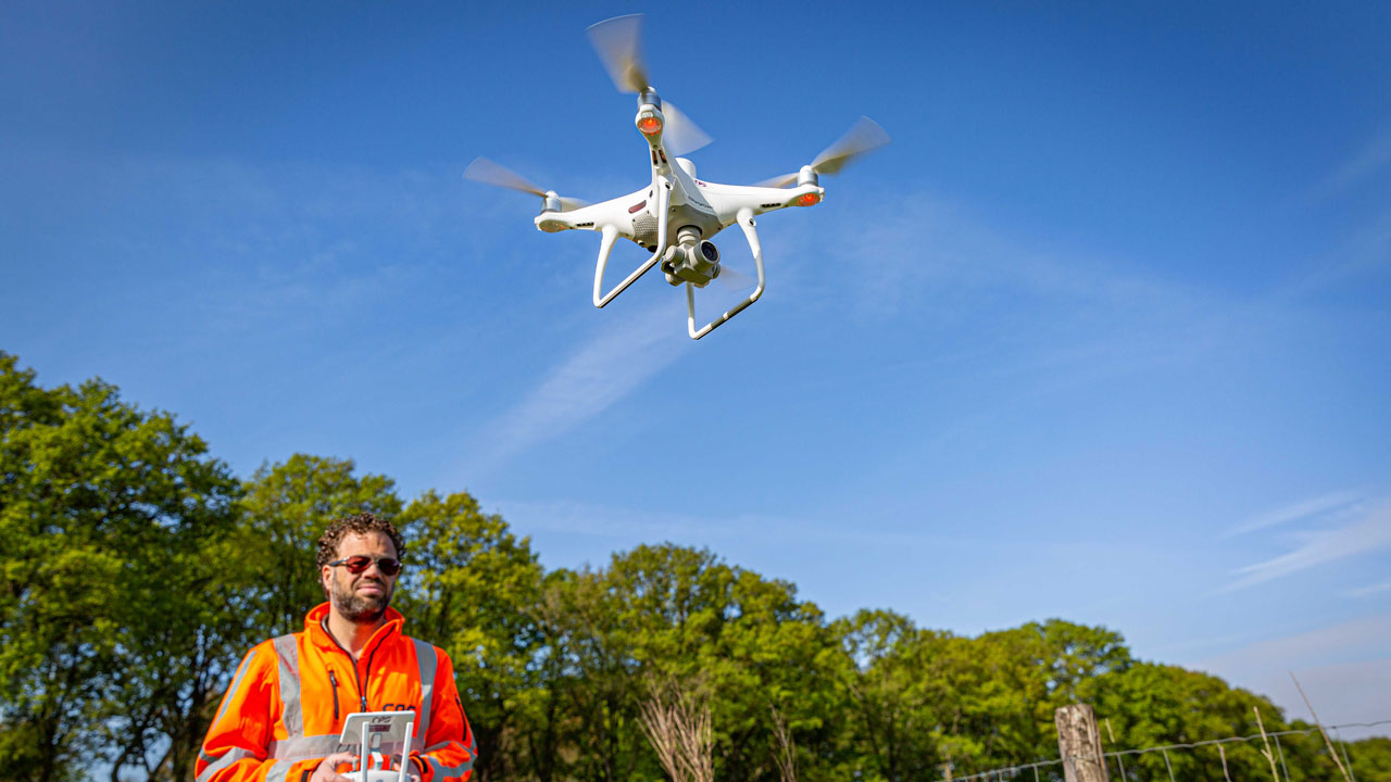 Dronepiloot in oranje veiligheidsjas bestuurt vliegende drone in buitengebied – visuele inspectie van infrastructuur met drone