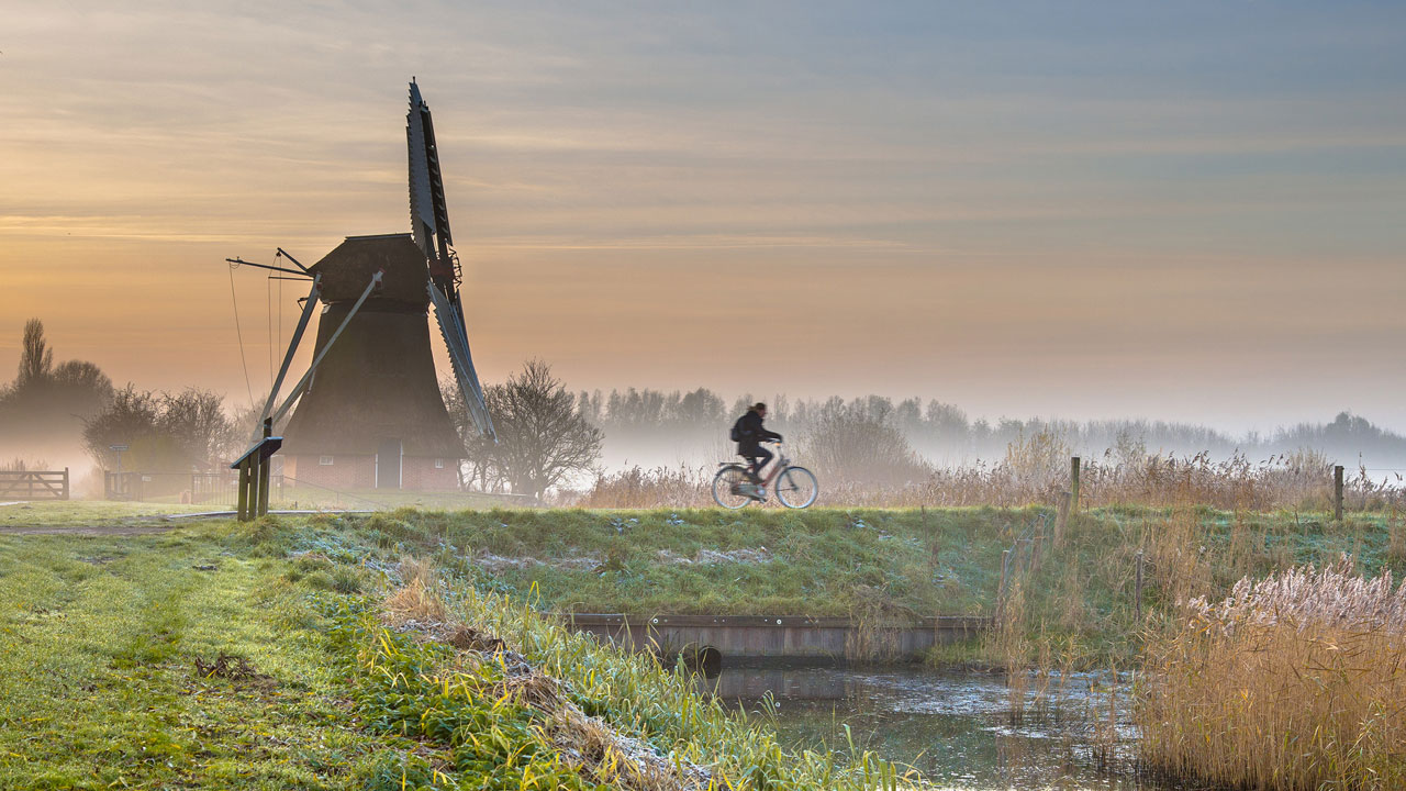 Fietser in Nederlands polderlandschap bij zonsopkomst met windmolen, mistige velden en water – symbool voor duurzame mobiliteit en milieubewust leven