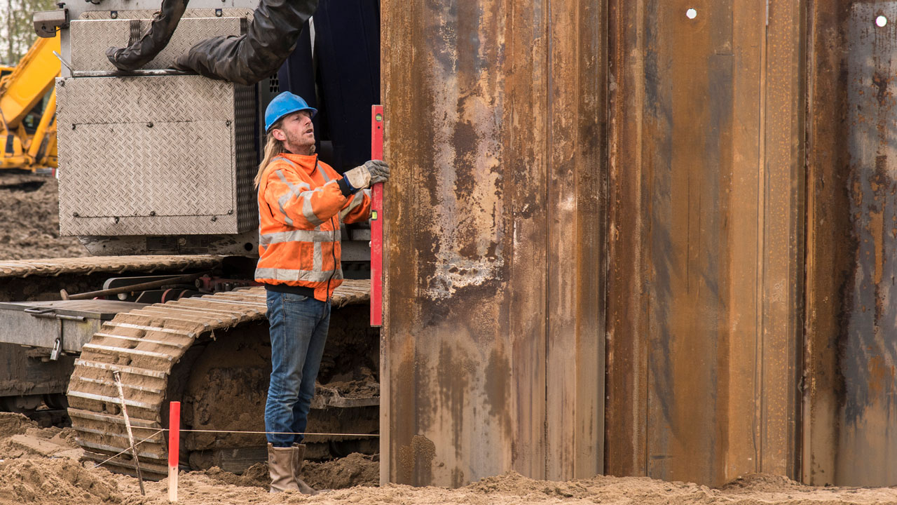 Bouwvakker meet stalen damwand met waterpas op zandige bouwlocatie, met geotechnisch materieel op de achtergrond