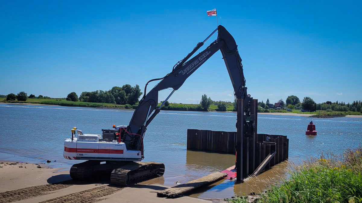 Graafmachine plaatst houten balk bij damwandconstructie langs rivier, met helder weer en groene oevers op de achtergrond