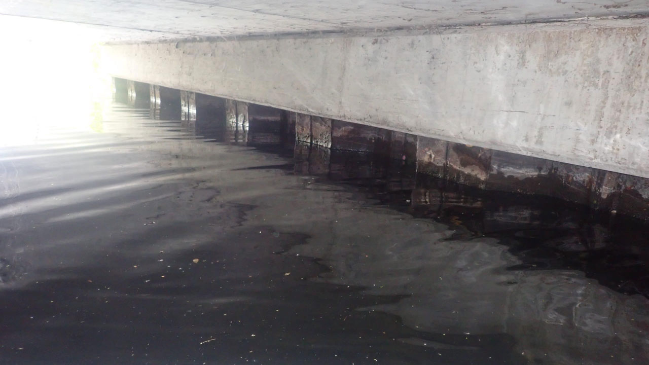 Onderzijde van betonnen brug met kalm stromend water en deels ondergedompelde steunpilaren, zichtbaar lichtinval van opzij