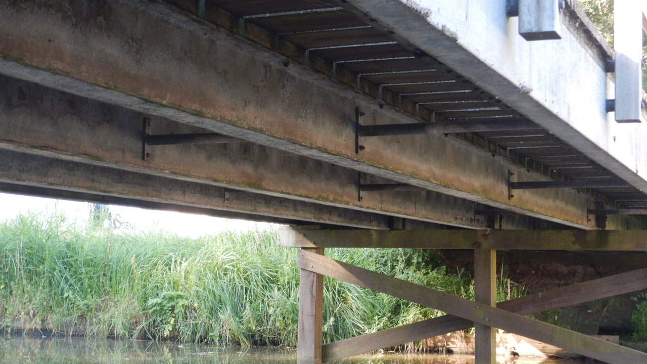 Onderzijde van brug met metalen liggers en houten palen boven water, zicht op constructieve details en reflectie in het water