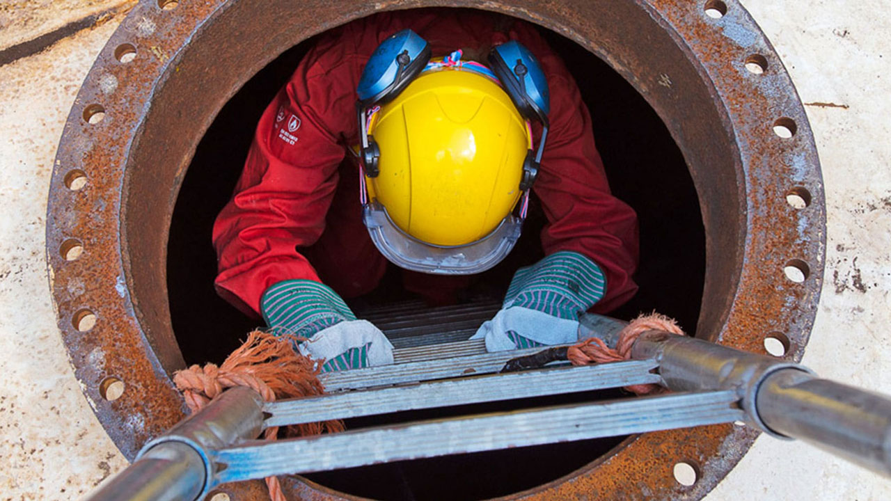 Inspecteur daalt via ladder af in mangat voor inspectie van besloten ruimte, met helm en beschermende kleding