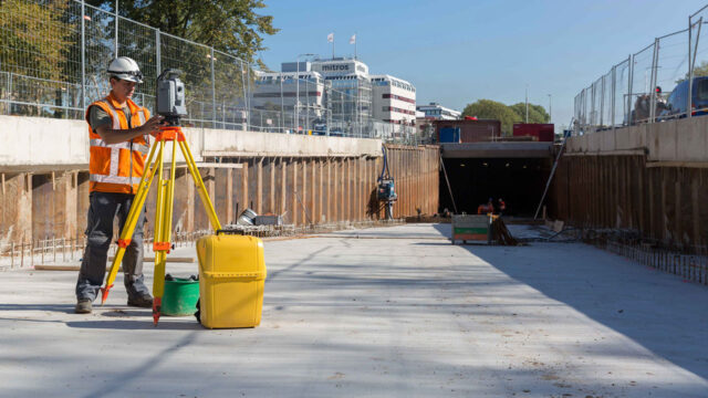 Inspecteur meet constructie in met total station op statief bij tunnelbouw, met zicht op betonwanden en achtergrondbebouwing