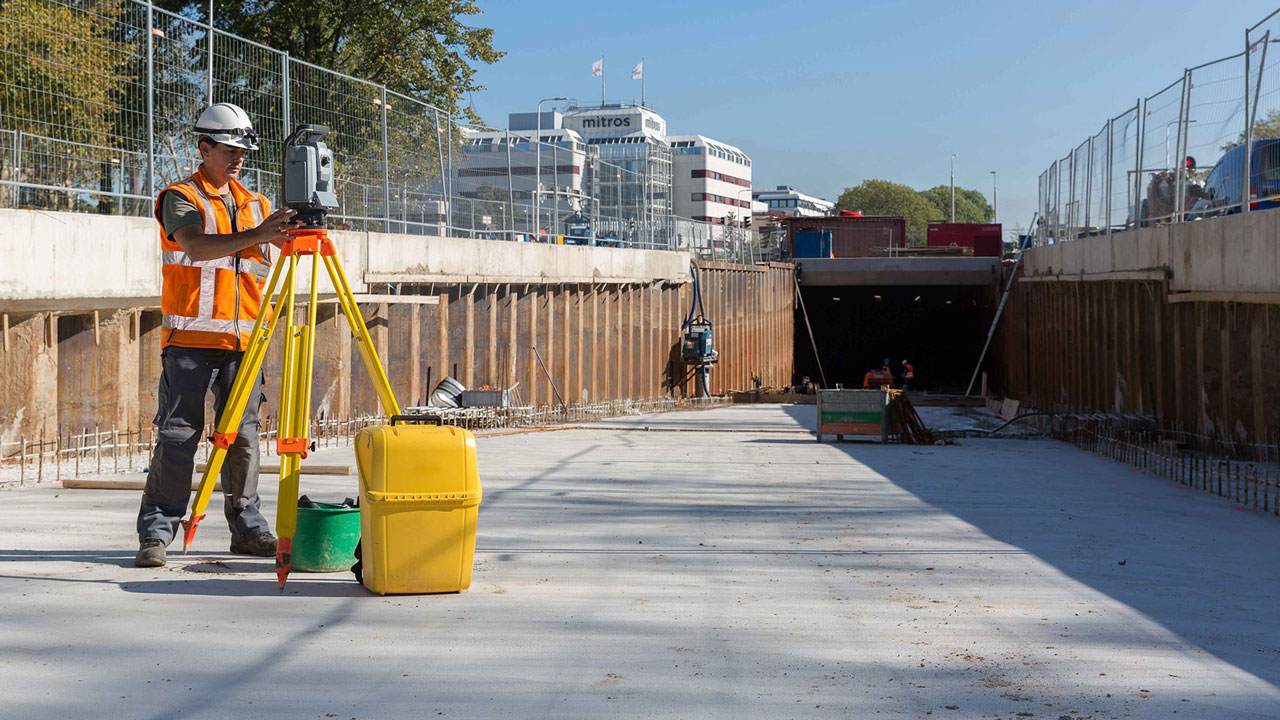 Inspecteur meet constructie in met total station op statief bij tunnelbouw, met zicht op betonwanden en achtergrondbebouwing