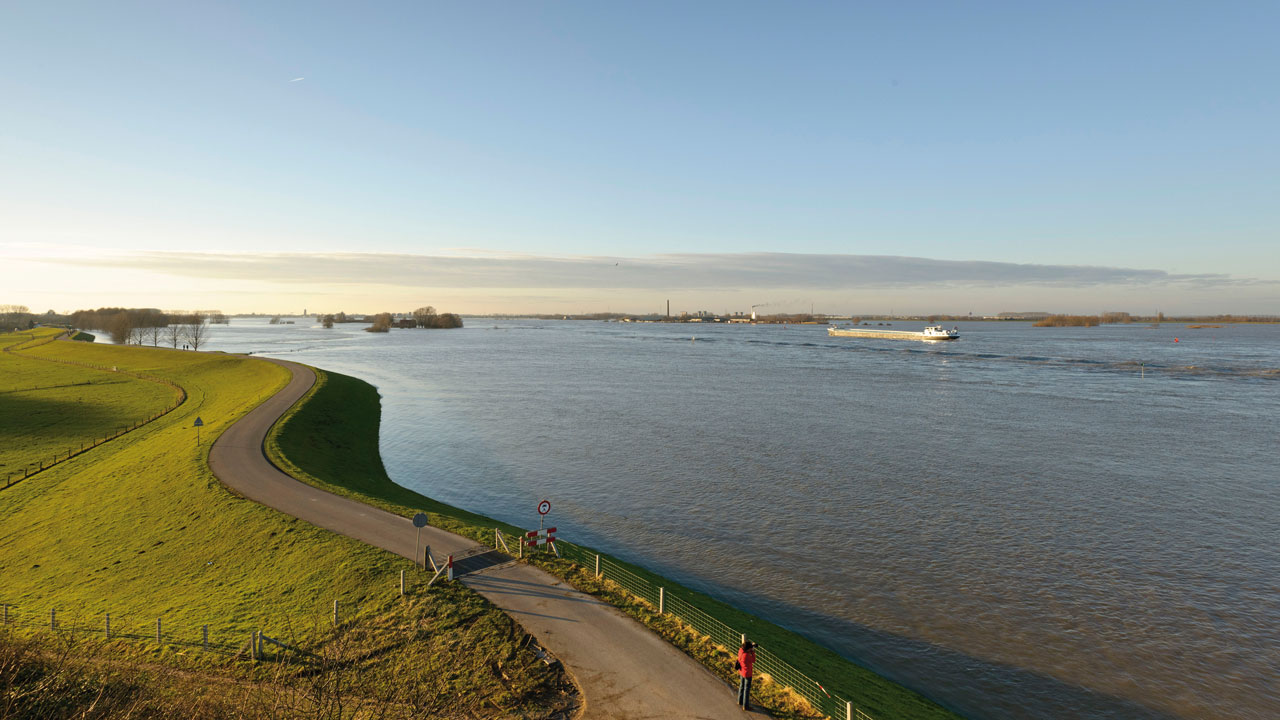 Brede rivier met wandelaars langs groene oever – balans tussen natuur, recreatie en infrastructuur in Nederlandse landschappen