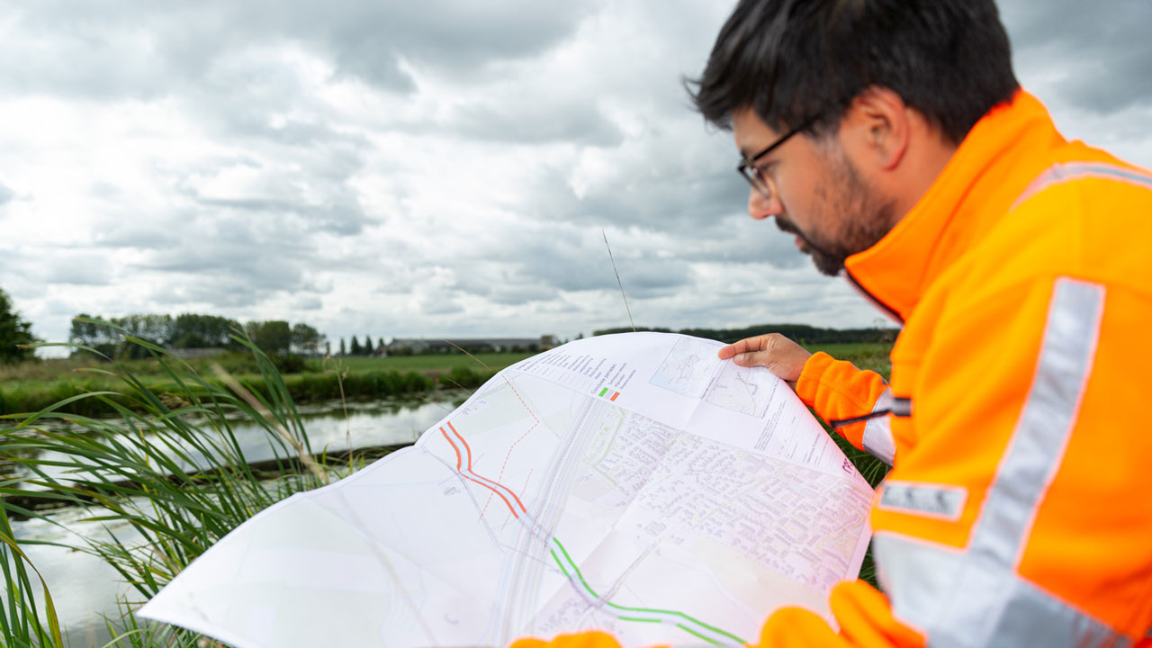Medewerker in oranje veiligheidsjas bekijkt kaart in buitengebied met water en groen – planning en uitvoering van natuurontwikkelingsprojecten
