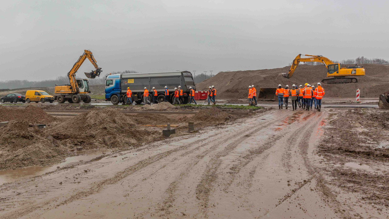 Actieve bouwplaats met graafmachines, vrachtwagens en medewerkers in oranje hesjes op modderig terrein.