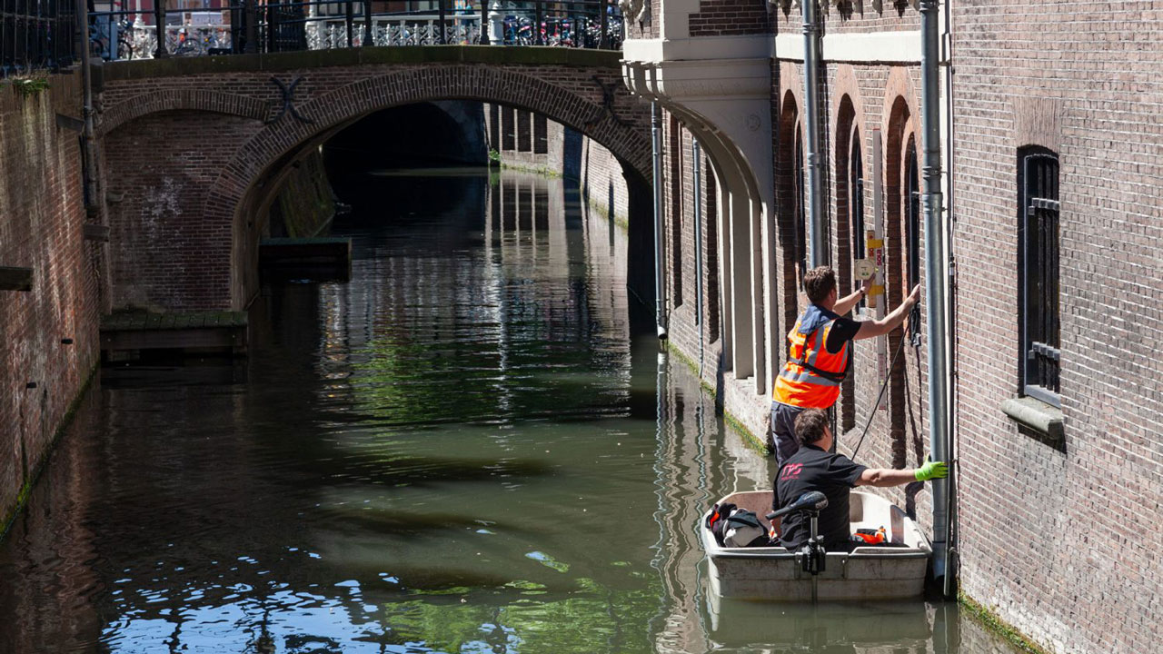 Inspecteurs in boot voeren meting uit aan kademuur in gracht, met zicht op bakstenen gevels en boogbrug op achtergrond