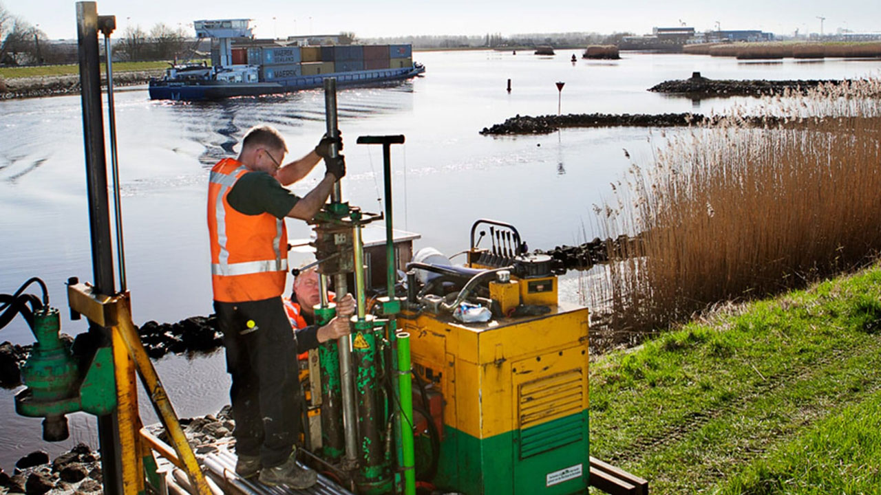 Medewerkers voeren grondonderzoek uit met boorinstallatie aan waterkant, met zicht op riet
