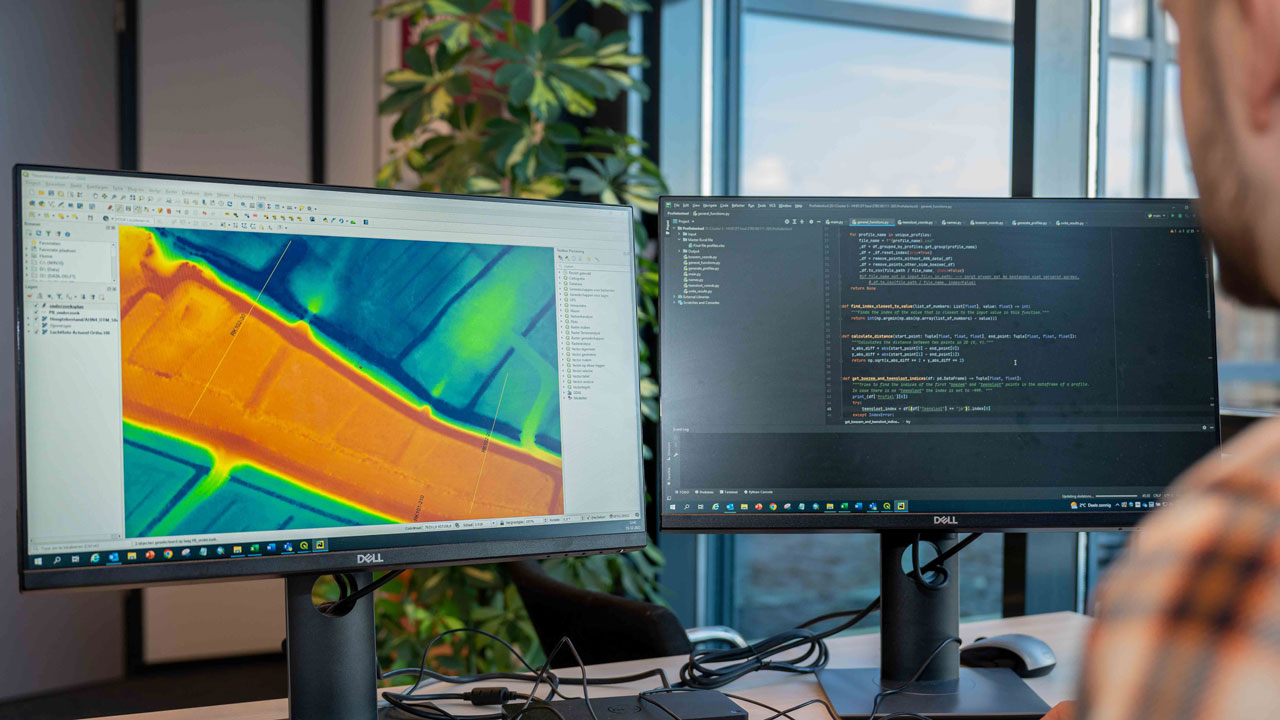 Een medewerker analyseert een topografische of thermische kaart op een computerscherm, met daarnaast een tweede scherm met code of data-analyse, gericht op waterveiligheid en infrastructuur