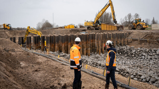 Twee medewerkers in oranje veiligheidskleding staan op een bouwplaats met graafmachines, een bulldozer en funderingspalen, omgeven door zand, grind en zware apparatuur