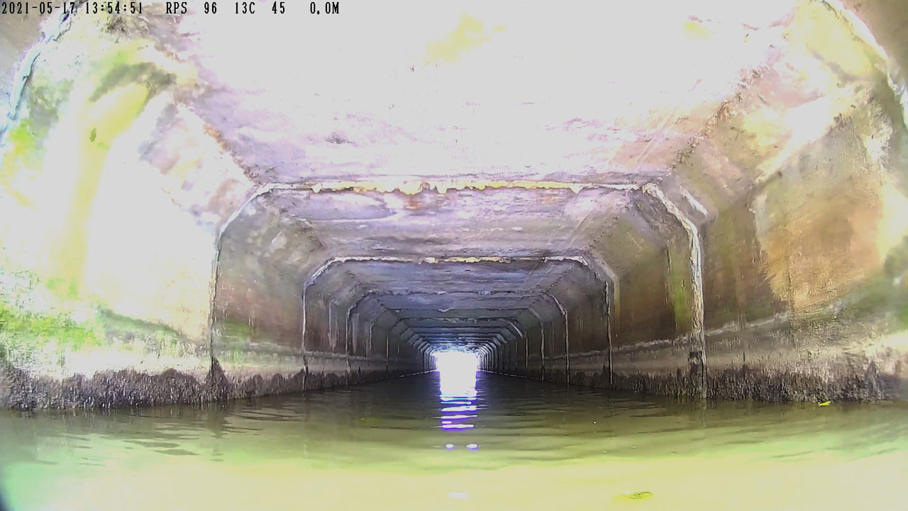 Binnenkant van betonnen tunnel met stromend water en lichtinval aan het einde, zichtbaar slijtage aan wanden en plafond