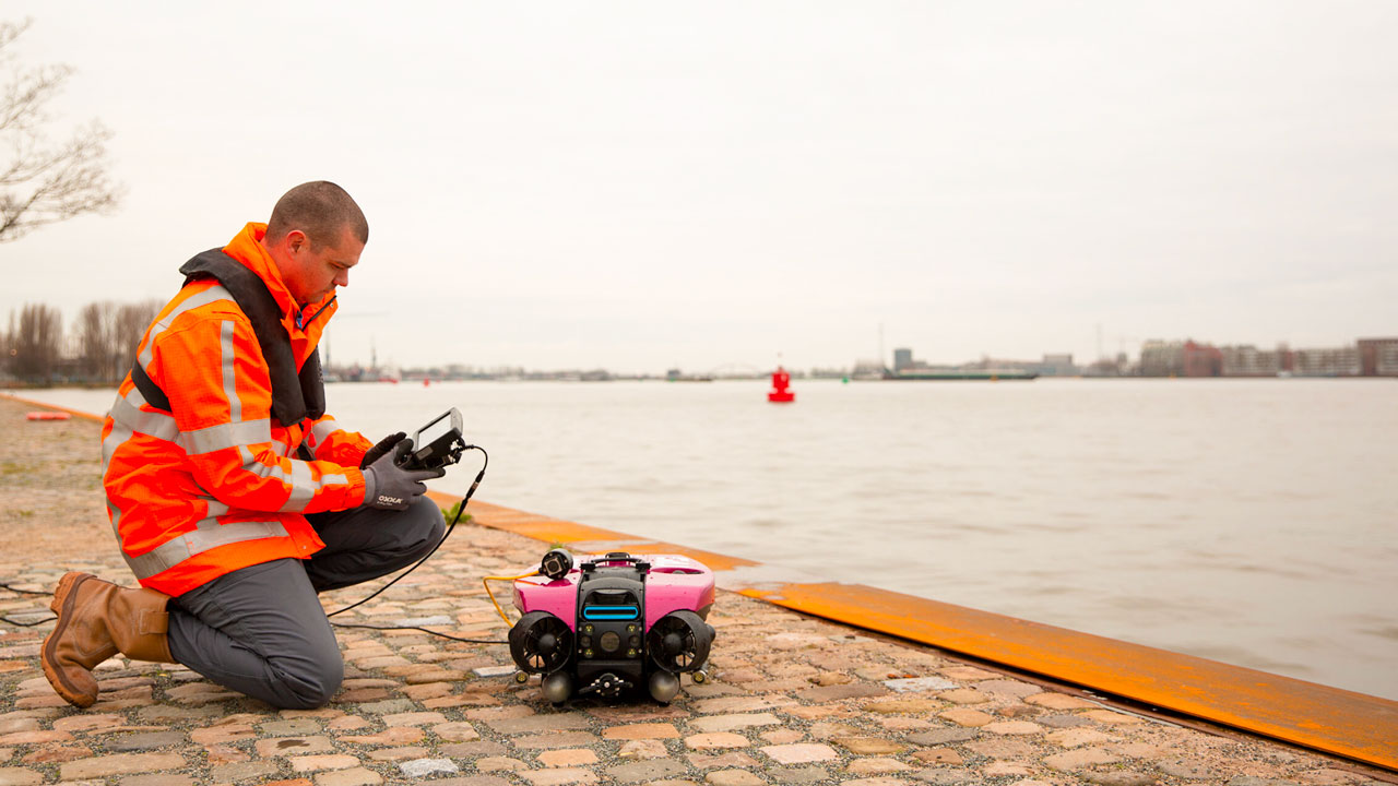 Inspecteur bedient roze ROV vanaf kade met afstandsbediening; water, boeien en gebouwen op achtergrond
