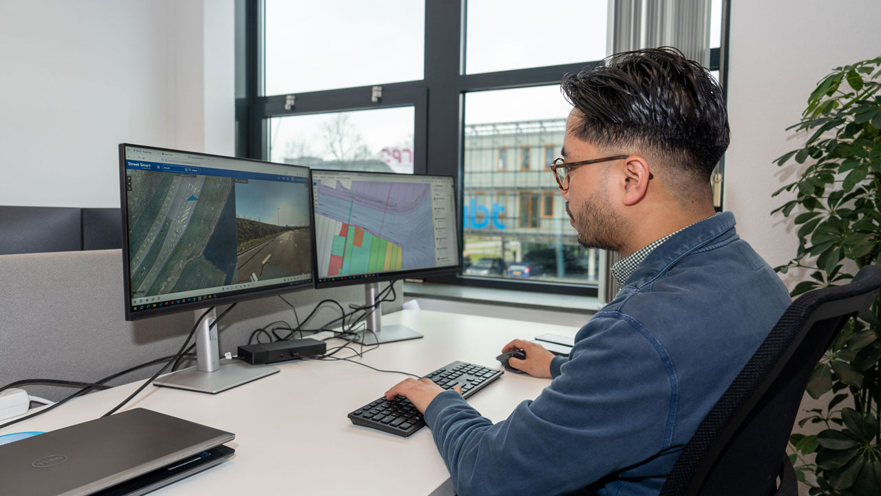 Medewerker werkt aan ontwerp- en kaartsoftware op meerdere schermen, in kantooromgeving met zicht op groen buitengebied