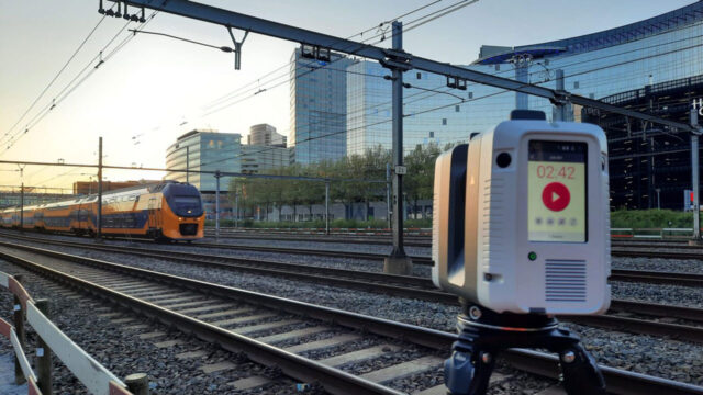 3D-laserscanner op statief bij spoorlijn met passerende trein en moderne gebouwen, bij zonsopkomst of zonsondergang