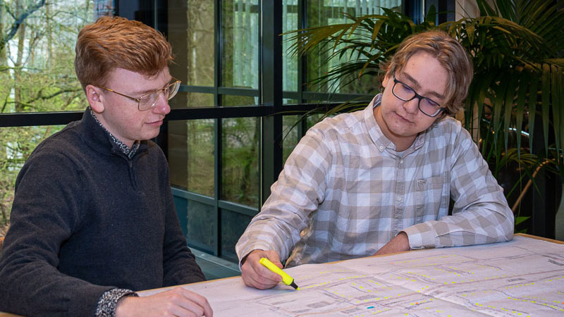 Twee mensen overleggen aan tafel met bouwtekening en markeerstift, met zicht op groen – focus op duurzame energietransitie