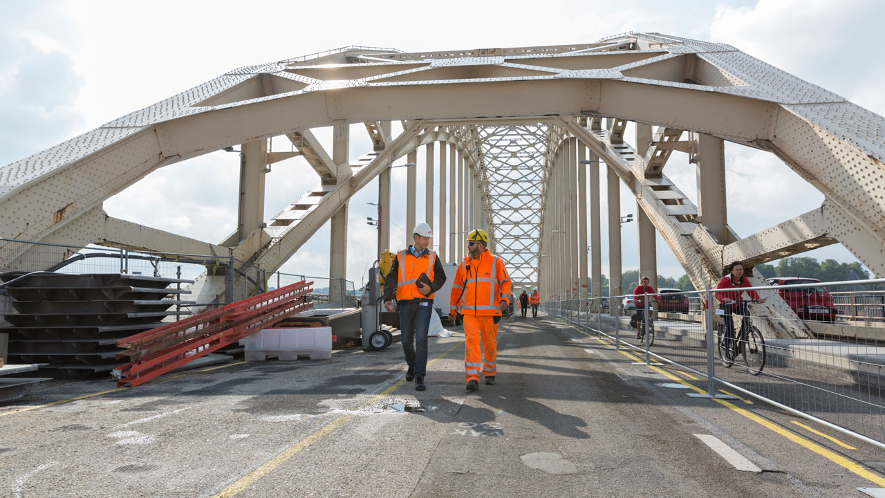 Onderhoudswerkzaamheden op brug met Tetra Tech-medewerkers en chroom-6-sanering, naast passerende fietsers en zichtbaar materieel