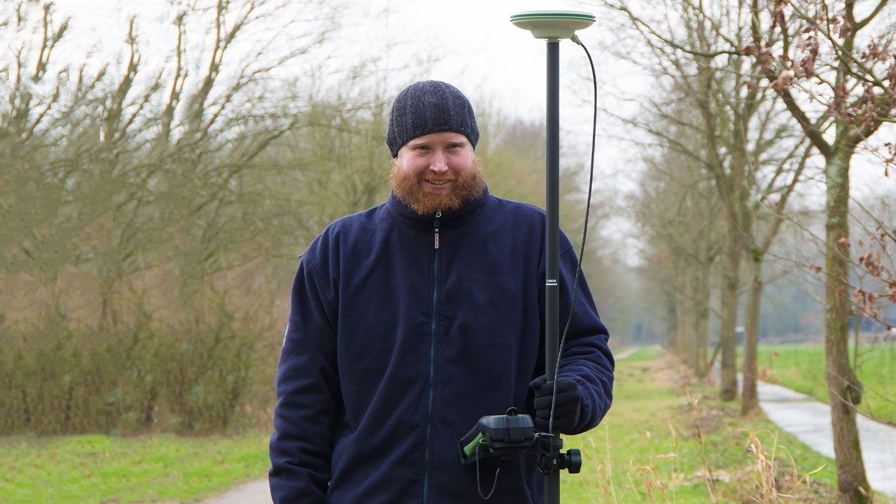 Medewerker voert landmeting uit in een natuurlijke omgeving met behulp van een meetinstrument. De achtergrond toont kale bomen en struiken, wat wijst op een herfst- of winterse setting