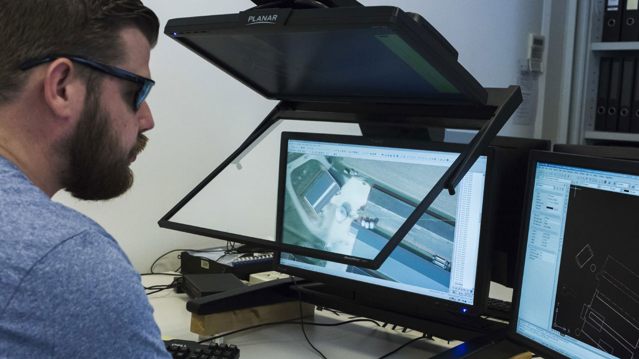 Medewerker werkt aan een bureau met meerdere computerschermen waarop technische tekeningen of ontwerpen zichtbaar zijn. Illustratie van digitale verwerking en analyse van meetgegevens