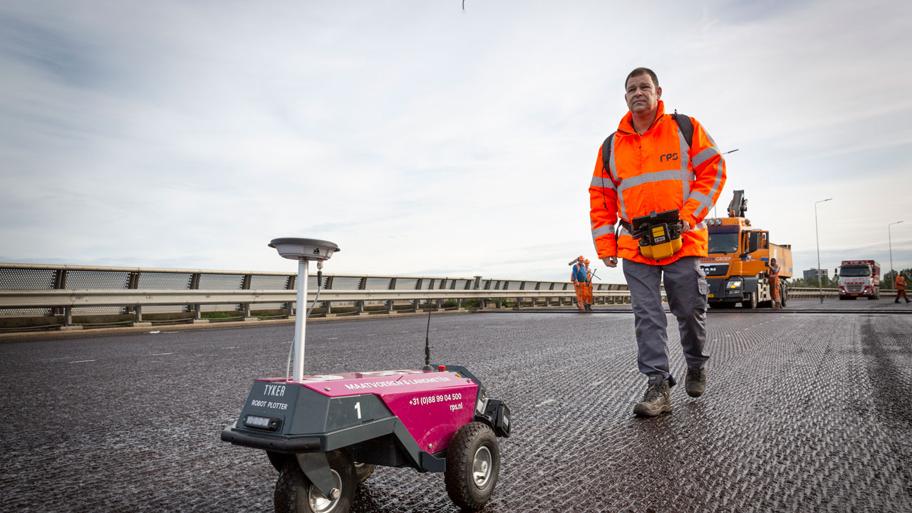 Persoon in oranje veiligheidskleding loopt naast een kleine, rijdende robot plotter. Op de achtergrond zijn andere medewerkers en voertuigen zichtbaar