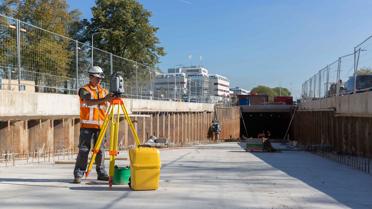 Landmeter met helm en oranje hesje gebruikt meetapparatuur op statief op bouwplaats met betonwanden, rebar en achtergrond met bomen en gebouwen