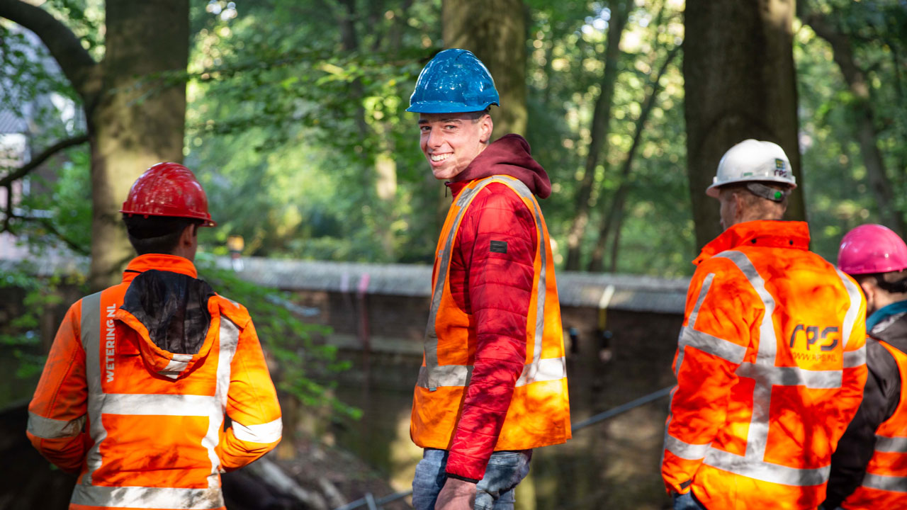 Studenten en experts in veiligheidskleding inspecteren renovatiewerkzaamheden boogbrug Naarden – samenwerking tussen generaties in techniek