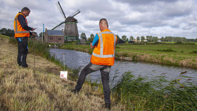 Tetra Tech-medewerkers meten bij watergang met windmolen op achtergrond: samenwerking in dijkversterking