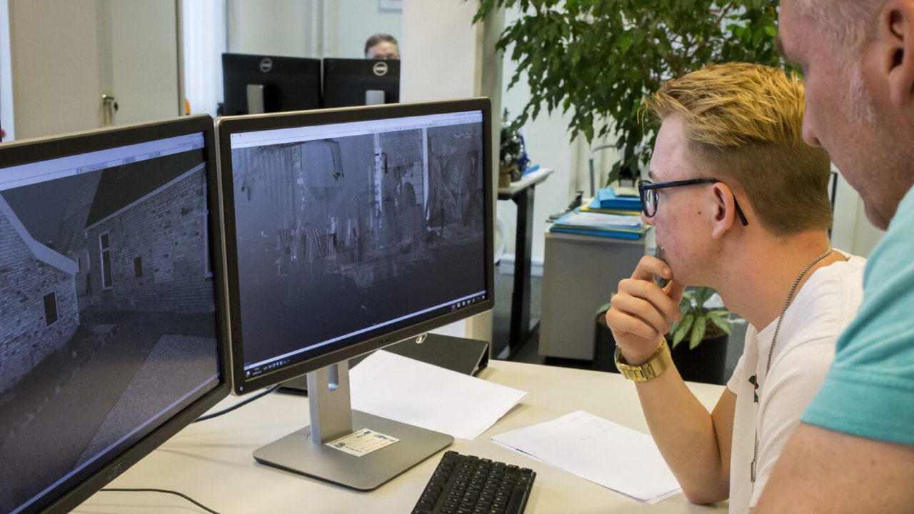 Twee personen bekijken computerschermen met topografische of bouwkundige beelden van gebouwen. Op het bureau liggen papieren en een toetsenbord. Illustratie van digitale verwerking van topografische meetgegevens